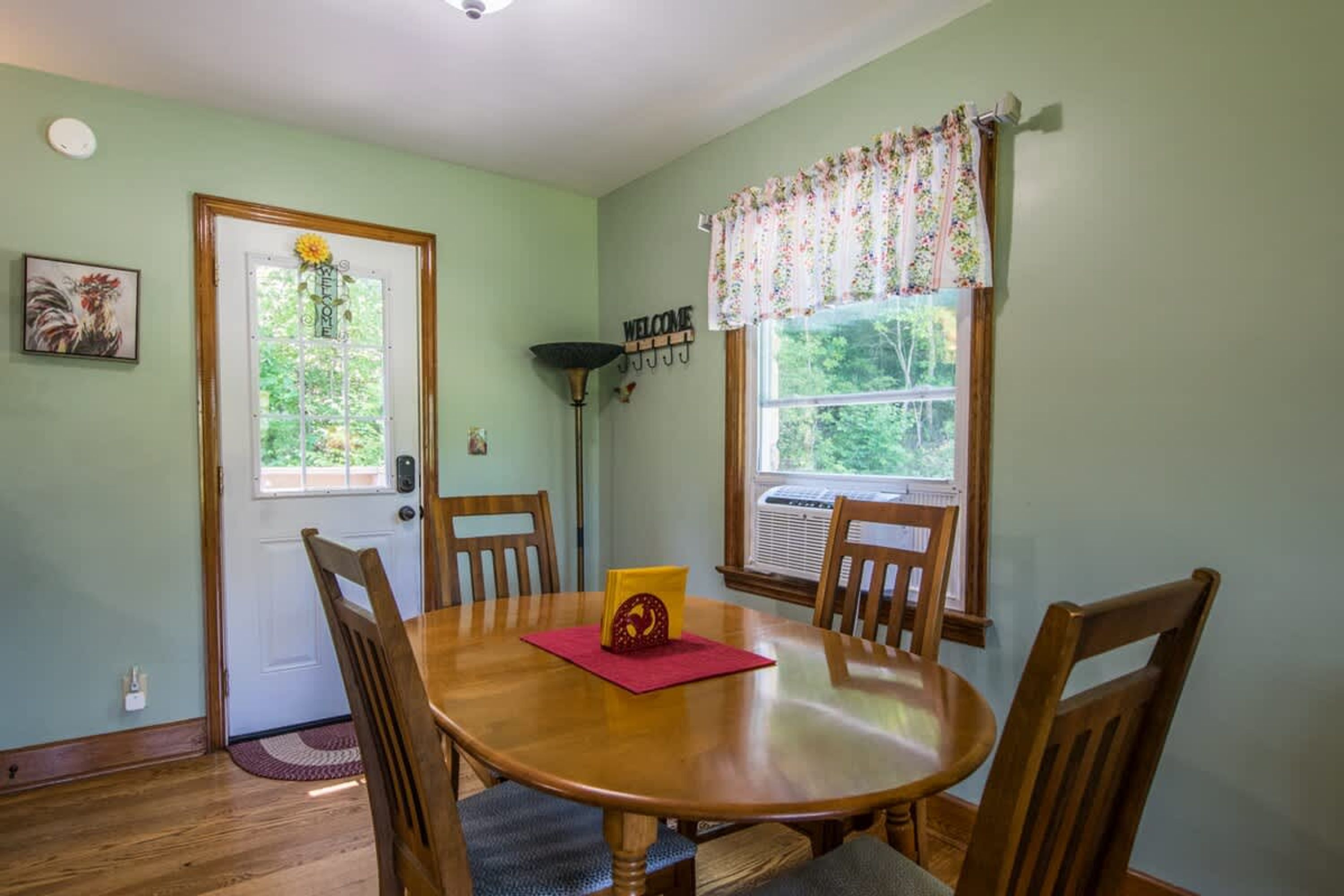 A dining area with a wooden table and four chairs, a door with a welcome sign, and a window with a floral curtain.
