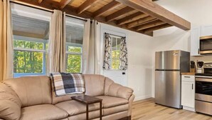 A cozy living room with a beige sofa, a small wooden table, and a patterned rug. There is a kitchen area with modern appliances, including a refrigerator and oven. - Accessible 1BR Cottage w Firepit Near Trails (Hendersonville)
