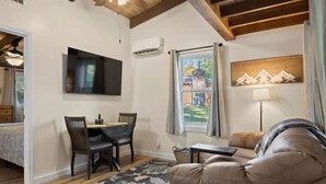 A cozy living room with a sofa, a dining table with chairs, a flat-screen TV mounted on the wall, and a window with curtains. - Accessible 1BR Cottage w Firepit Near Trails (Hendersonville)