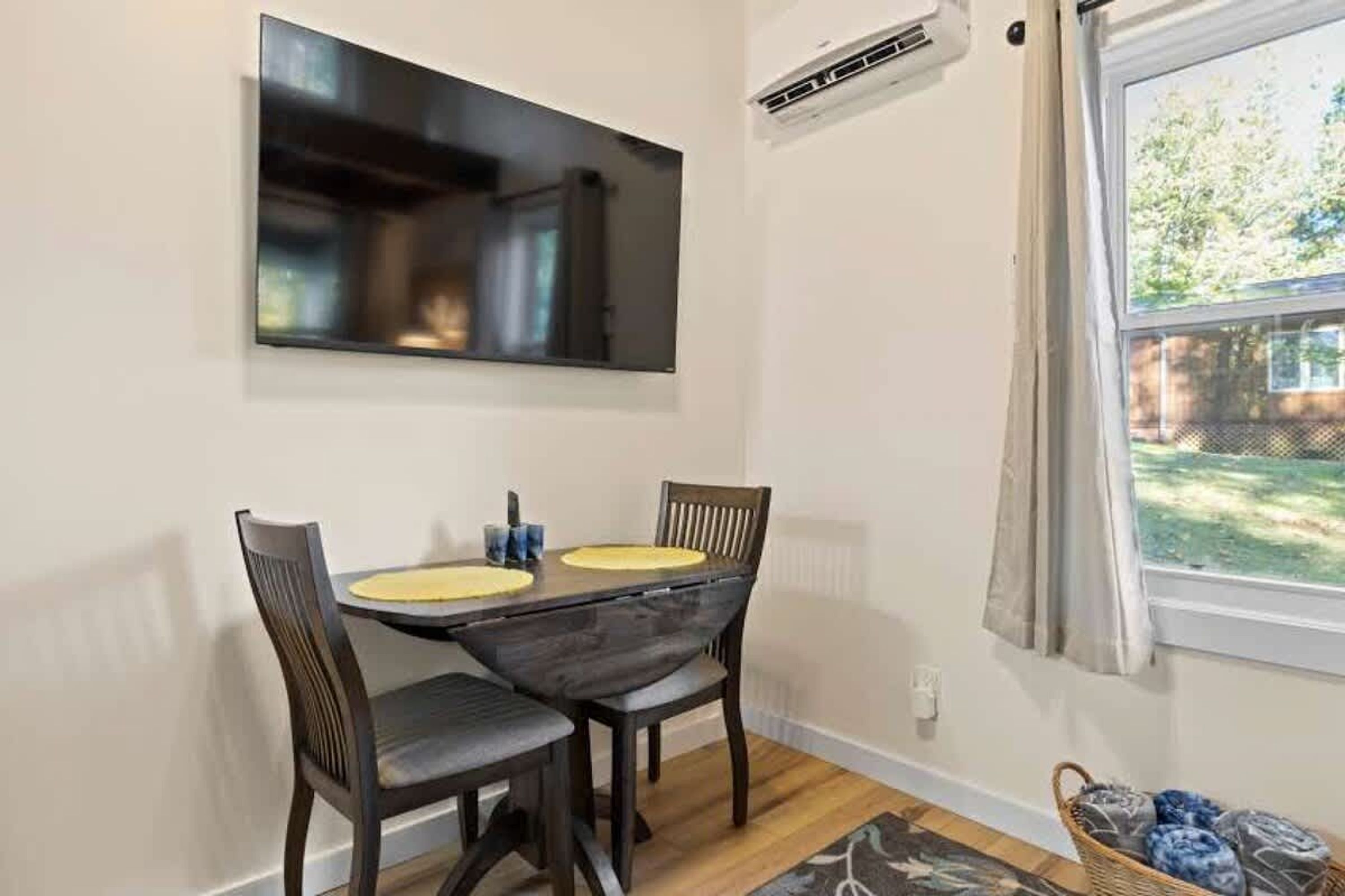 Un comedor con una mesa y sillas de madera oscura, un televisor de pantalla plana montado arriba y una ventana con cortinas.