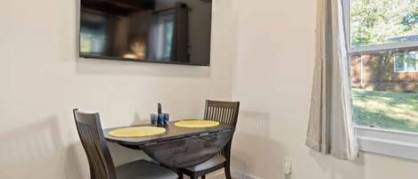 Un comedor con una mesa y sillas de madera oscura, un televisor de pantalla plana montado arriba y una ventana con cortinas.