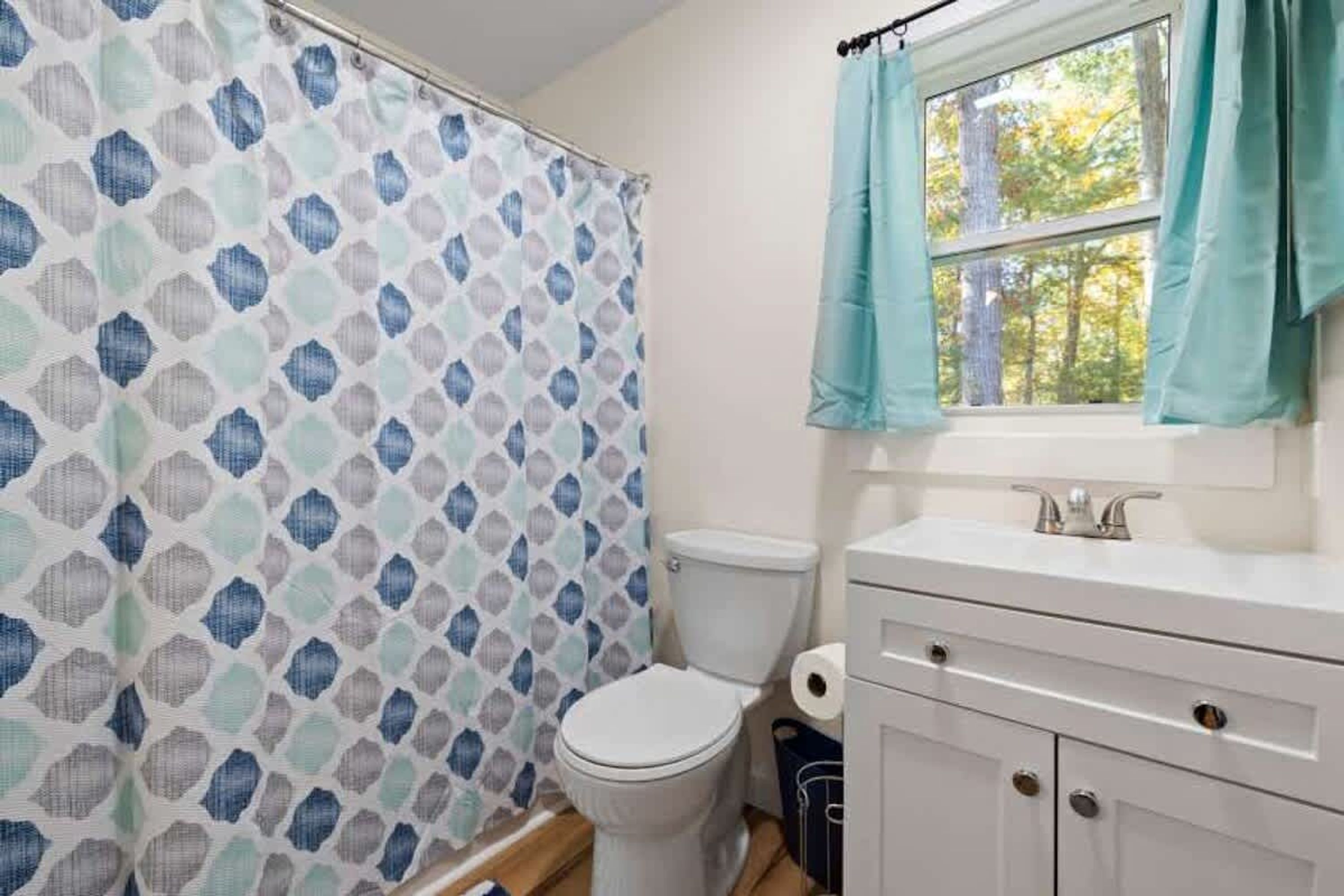 Une salle de bain avec un rideau de douche à motifs, des toilettes, un lavabo avec un miroir et une fenêtre avec des rideaux.