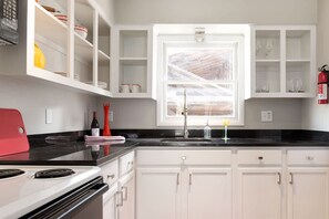 A kitchen with white cabinets, a black countertop, a red appliance, and a window with a view of a wooden structure. - Cheerful Studio w Patio Steps From Everything (Asheville)