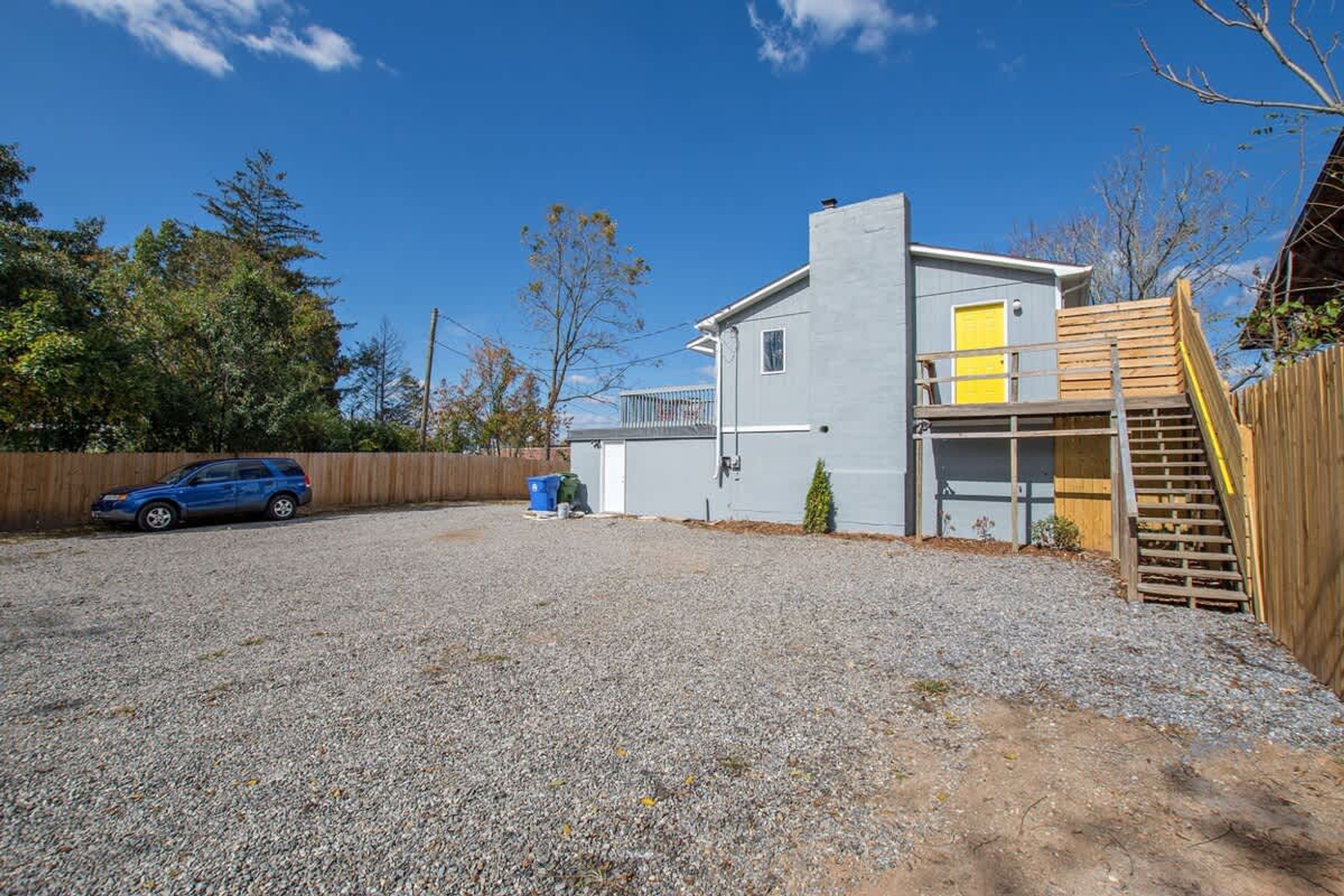 Une maison moderne à deux étages, avec une allée en gravier, une voiture bleue garée sur le côté gauche et une clôture en bois sur le côté droit.