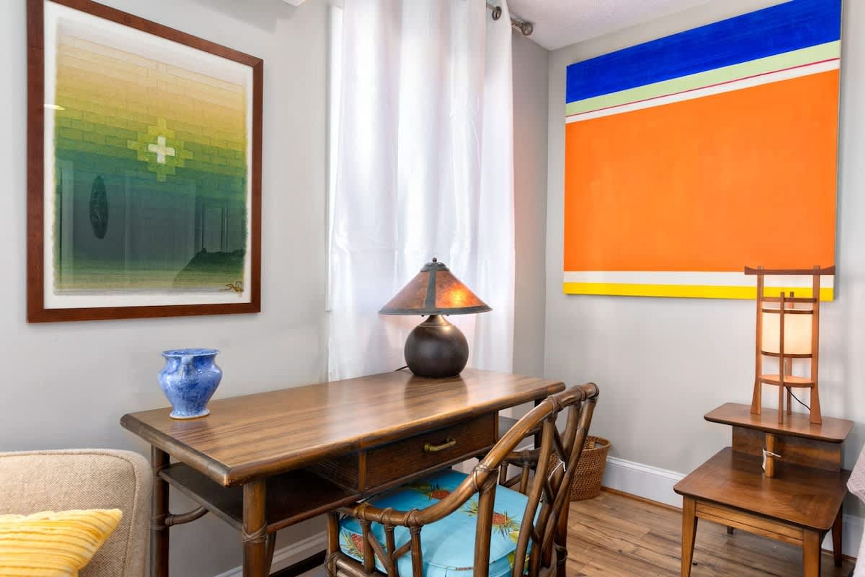 A room with a wooden desk, a lamp, a chair, a blue vase, and a framed picture on the wall.
