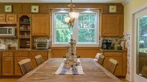 A kitchen with wooden cabinets, a dining table with chairs, a chandelier, and a window with a view of greenery. - Historic 4BR Colonial w Pool Table Fire Pit (Hendersonville)