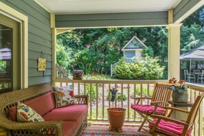 A porch with a wicker sofa and a wooden chair, a potted plant, and a view of a house surrounded by greenery. - Historic 4BR Colonial w Pool Table Fire Pit (Hendersonville)