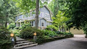 A two-story house with a front porch, surrounded by lush greenery and stone steps leading to the entrance. - Historic 4BR Colonial w Pool Table Fire Pit (Hendersonville)