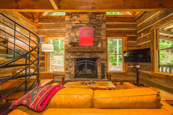 A log cabin interior with a stone fireplace, a sofa, a TV, and a spiral staircase. - Epic 1BR Cabin w Outdoor Fireplace 5 Min to Town (Saluda)