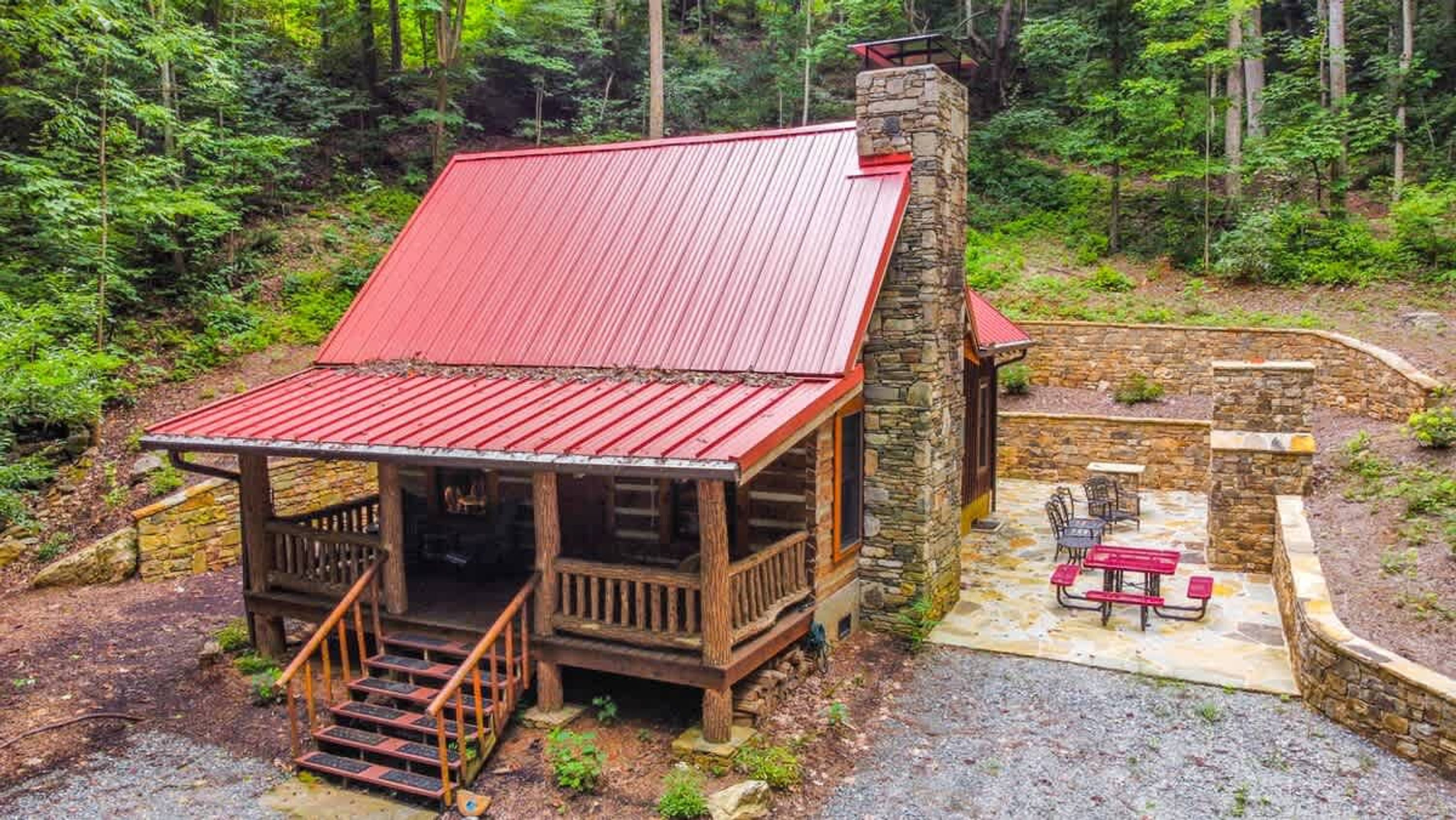 A cabin with a red roof, stone chimney, and a porch with steps leading to a wooden door.