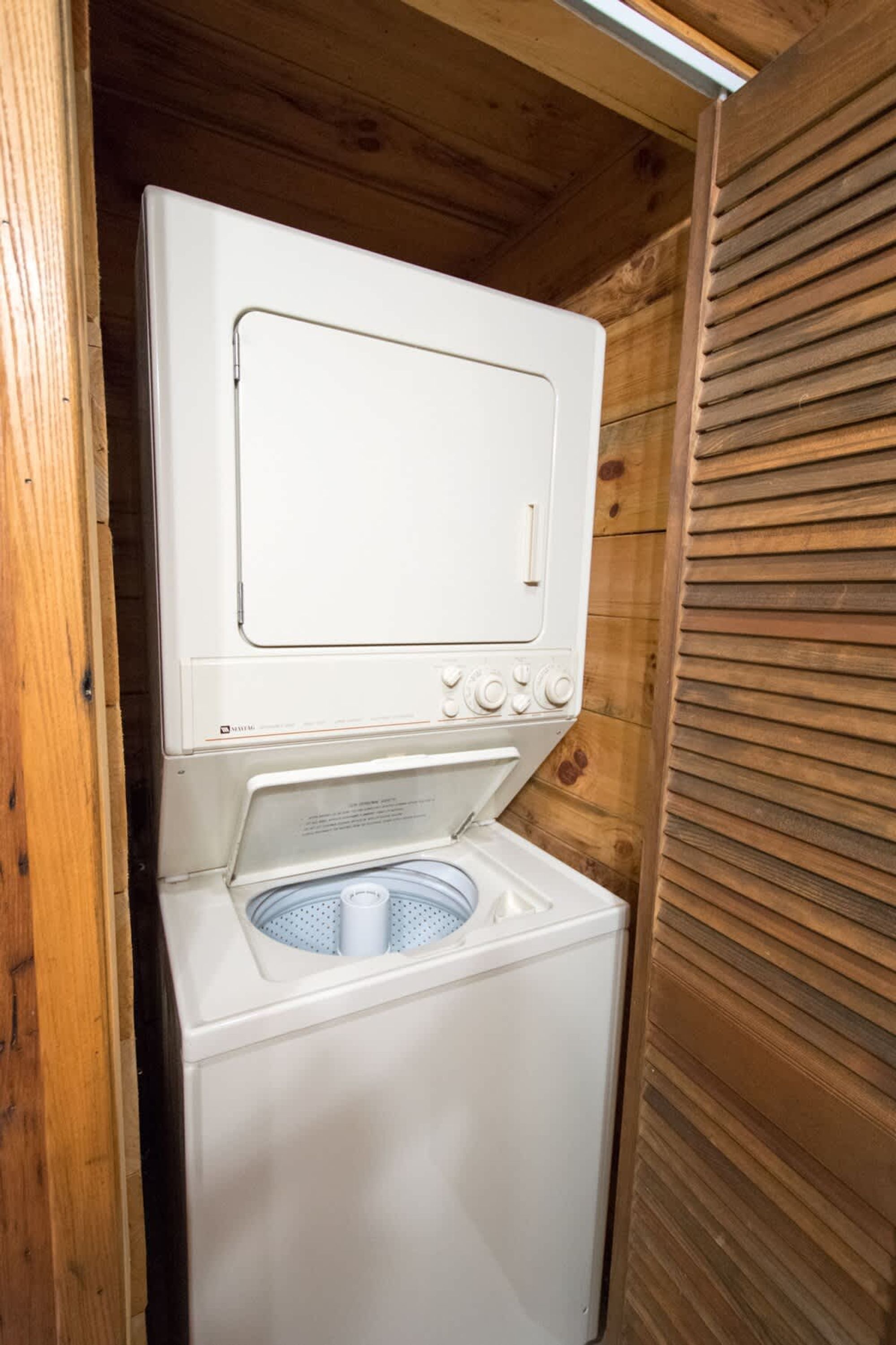 A compact, white, wall-mounted washer and dryer unit with a front-loading washing machine and a top-loading dryer, enclosed in a wooden cabin-style room.