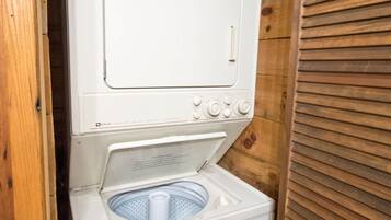 A compact, white, wall-mounted washer and dryer unit with a front-loading washing machine and a top-loading dryer, enclosed in a wooden cabin-style room.