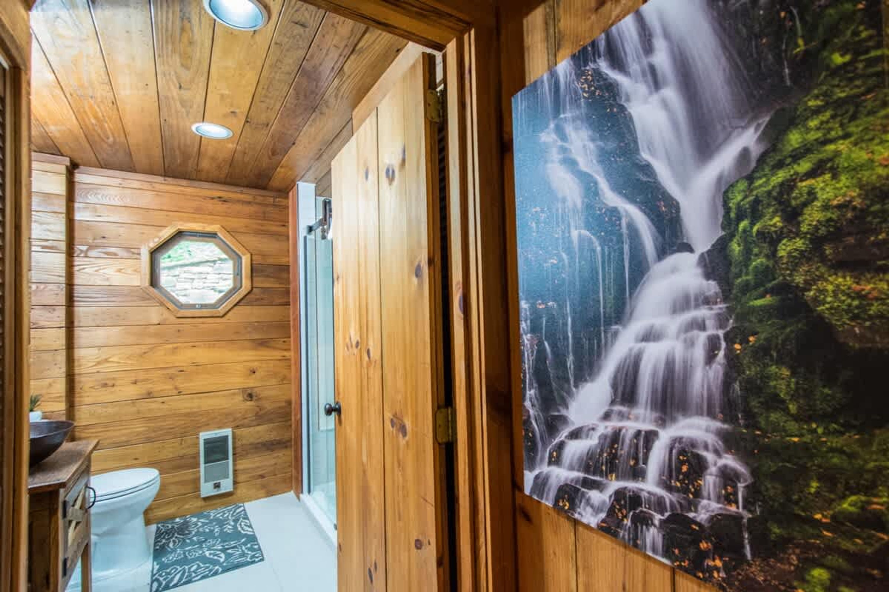 A wooden interior with a shower, a toilet, and a large waterfall mural.
