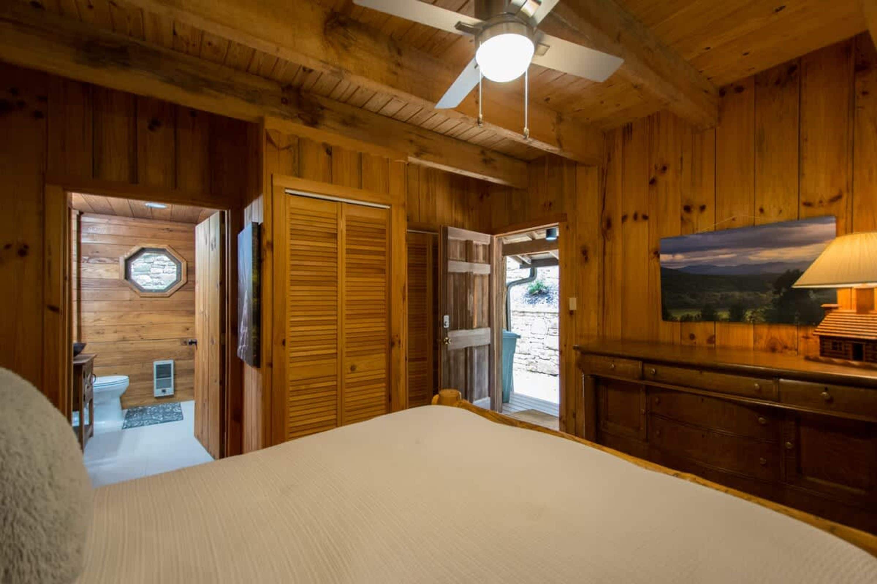 Un dormitorio de cabaña de madera con una cama, un ventilador de techo, un cuadro y vistas al exterior.