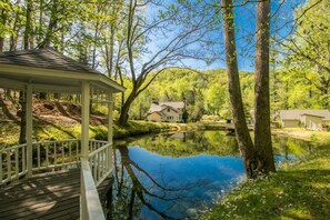 A gazebo with a wooden deck overlooking a calm pond, surrounded by lush greenery and a house in the background. - Rustic 3BR w Private Pond Firepit Near Trails (Hendersonville)