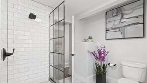 A modern bathroom with white subway tiles, a glass shower enclosure, a white toilet, and a black abstract painting on the wall. - Modern 1BR w Workspace Walk to Shops Parks (Asheville)