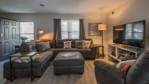 A modern living room with a sectional sofa, a coffee table, a TV, and a lamp. - Charming 2BR Townhouse w Patio Walk to Main St (Hendersonville)