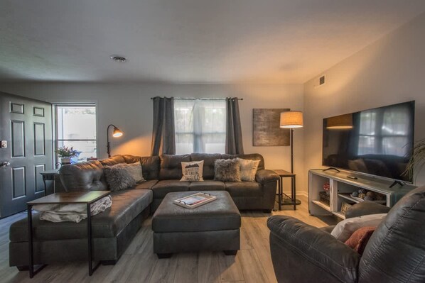 A modern living room with a sectional sofa, a coffee table, a TV, and a lamp. - Charming 2BR Townhouse w Patio Walk to Main St (Hendersonville)