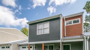 A two-story residential building with a garage, a covered porch, and a well-maintained lawn. - Charming 2BR Townhouse w Patio Walk to Main St (Hendersonville)