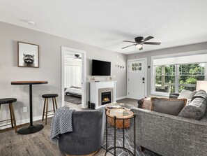 A modern living room with a fireplace, a television, and a large window offering a view of the outdoors. - Spacious 3BR Retreat w Game Room Near Pisgah (Candler)