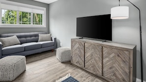 Ein modernes Wohnzimmer mit grauem Sofa, einem Flachbildfernseher auf einem Holzschrank und einem großen Fenster mit Blick auf Bäume.