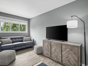 A modern living room with a grey sofa, a flat-screen TV on a wooden cabinet, and a large window with a view of trees. - Spacious 3BR Retreat w Game Room Near Pisgah (Candler)