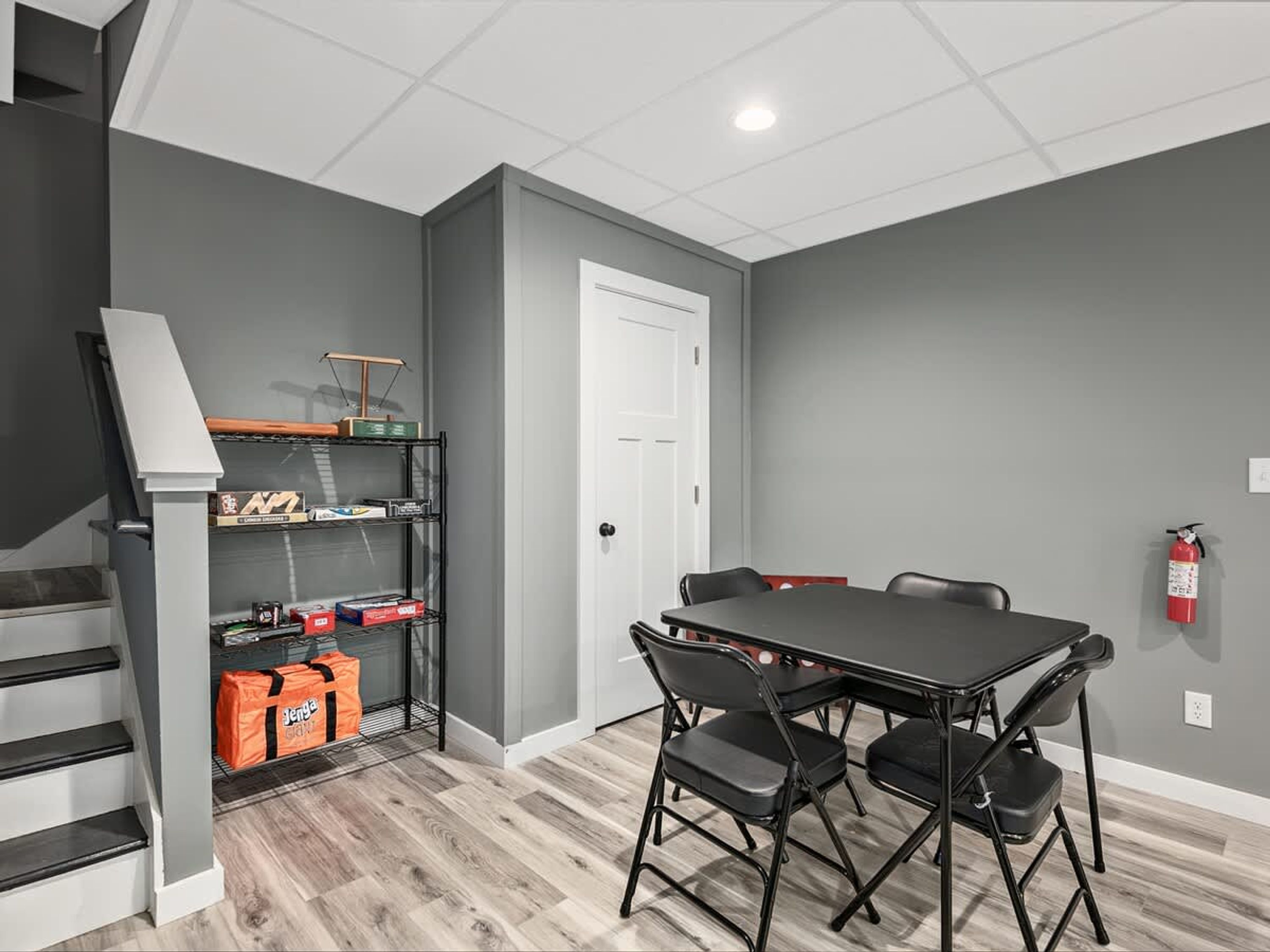 A room with a dining table and chairs, a staircase, a white door, and a shelf with boxes and a bag.