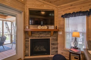 A cozy cabin interior with a stone fireplace, a flat-screen TV mounted above it, and a window with blinds. - Lakeview 2BR Tiny Home Near Trails Pool Bocce (Flat Rock)
