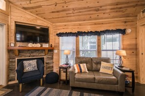 A cozy cabin interior with a stone fireplace, a leather sofa with a 'sweet' pillow, a blue armchair, and a television mounted on the wall. - Lakeview 2BR Tiny Home Near Trails Pool Bocce (Flat Rock)