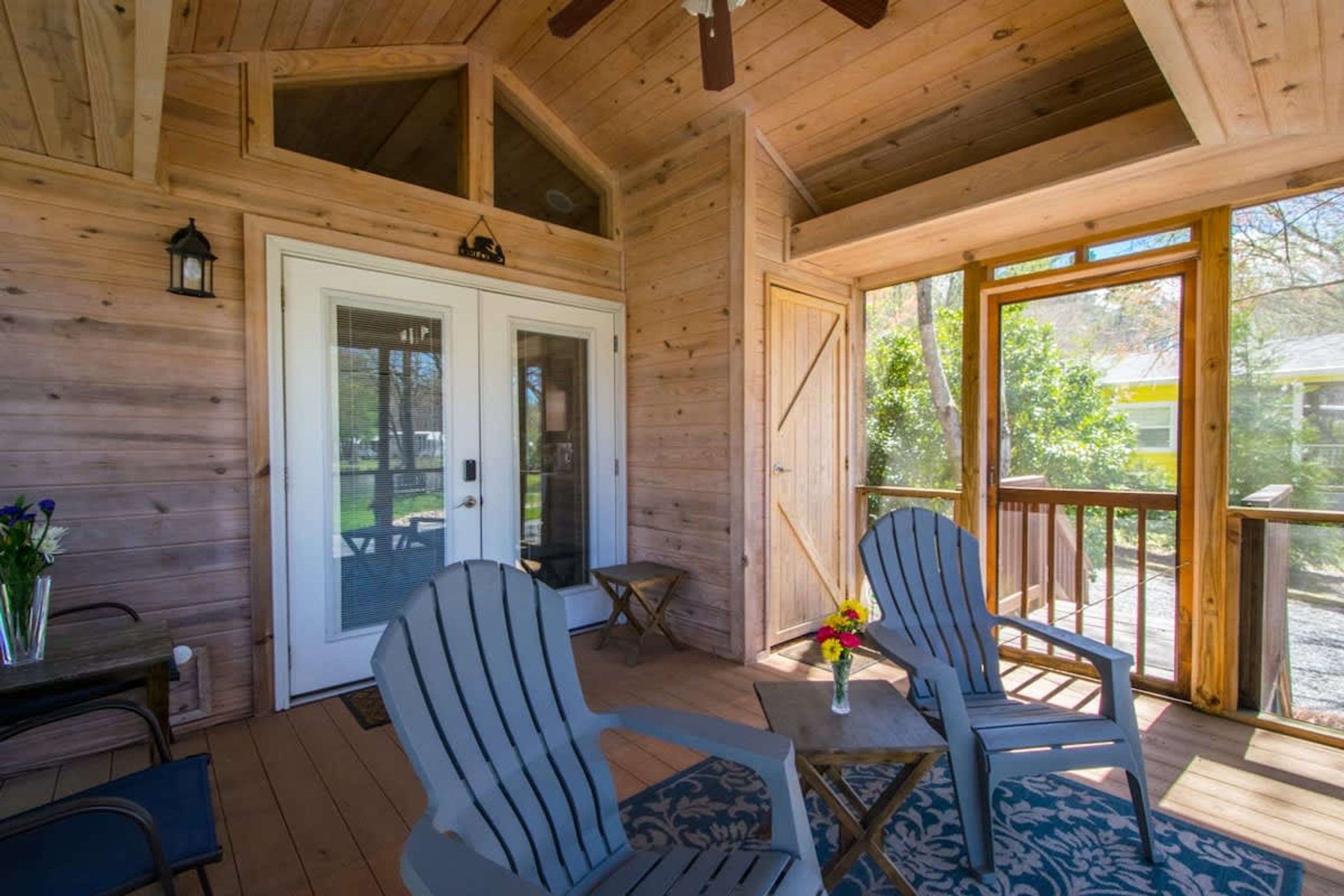 A wooden cabin interior with two Adirondack chairs, a table with a vase of flowers, and a sliding glass door leading outside.