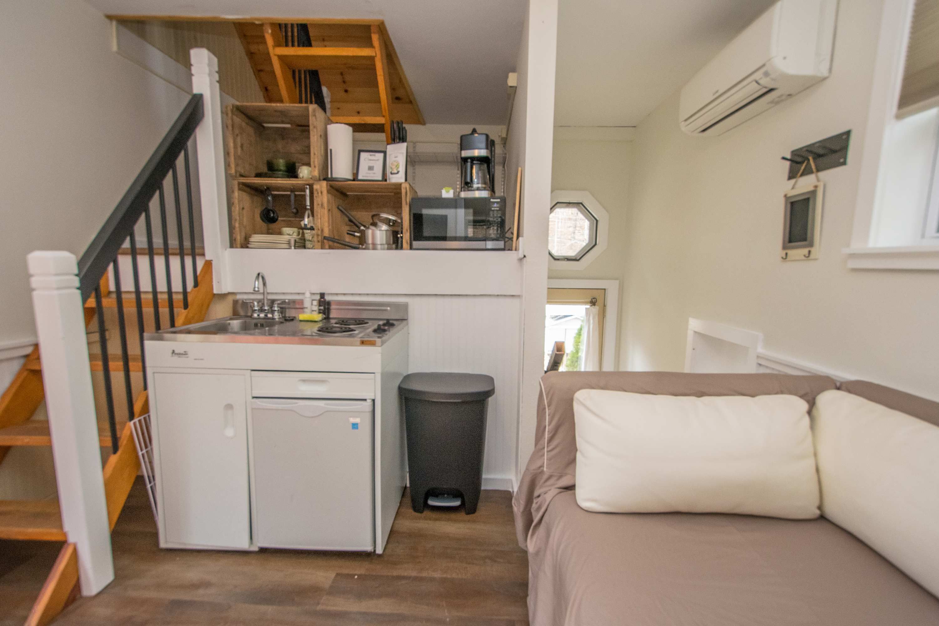 A compact kitchen with a stove, sink, and coffee maker, a small trash bin, and a staircase leading to a loft area.