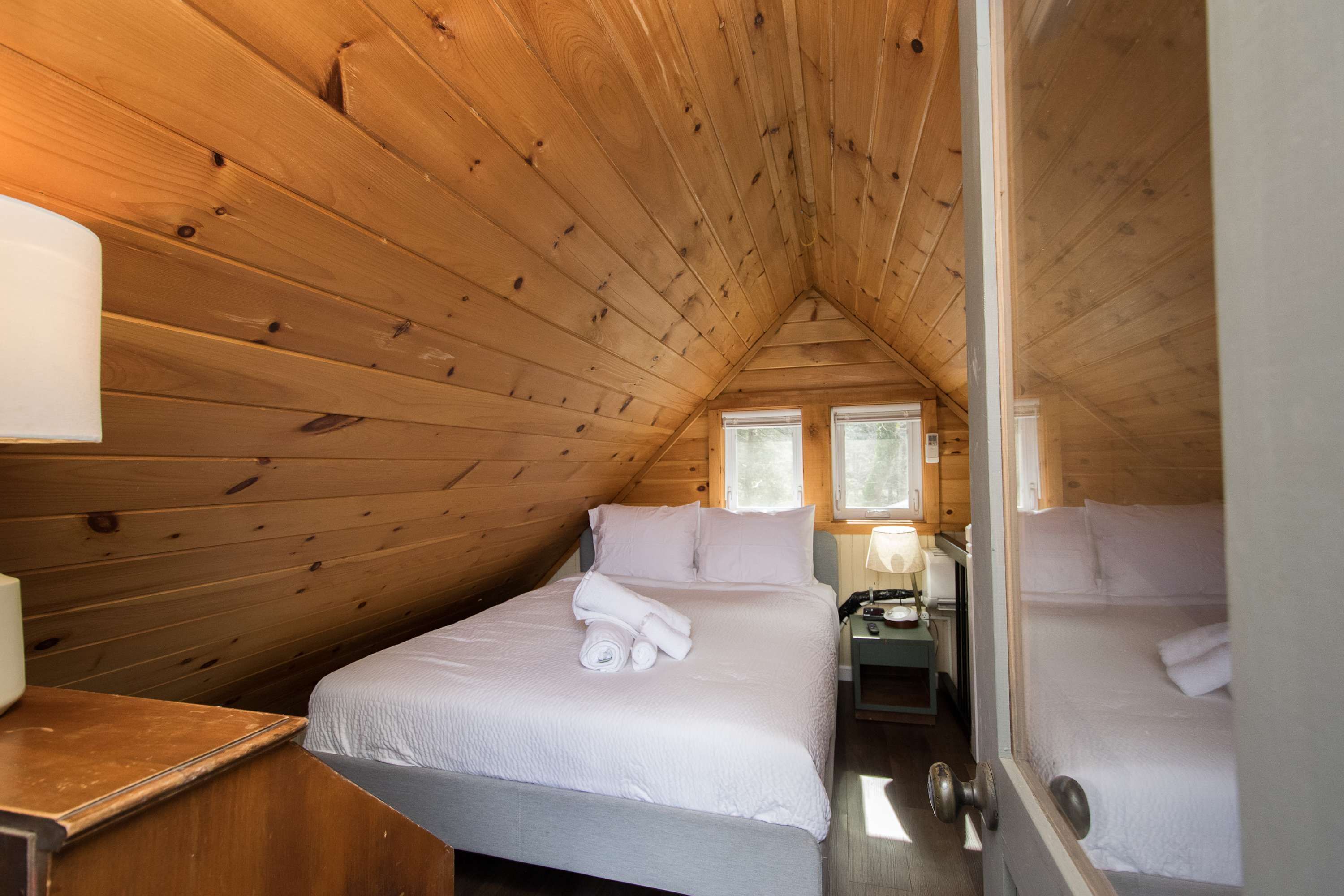 A cozy attic bedroom with wooden walls, two beds, a nightstand, and a window.