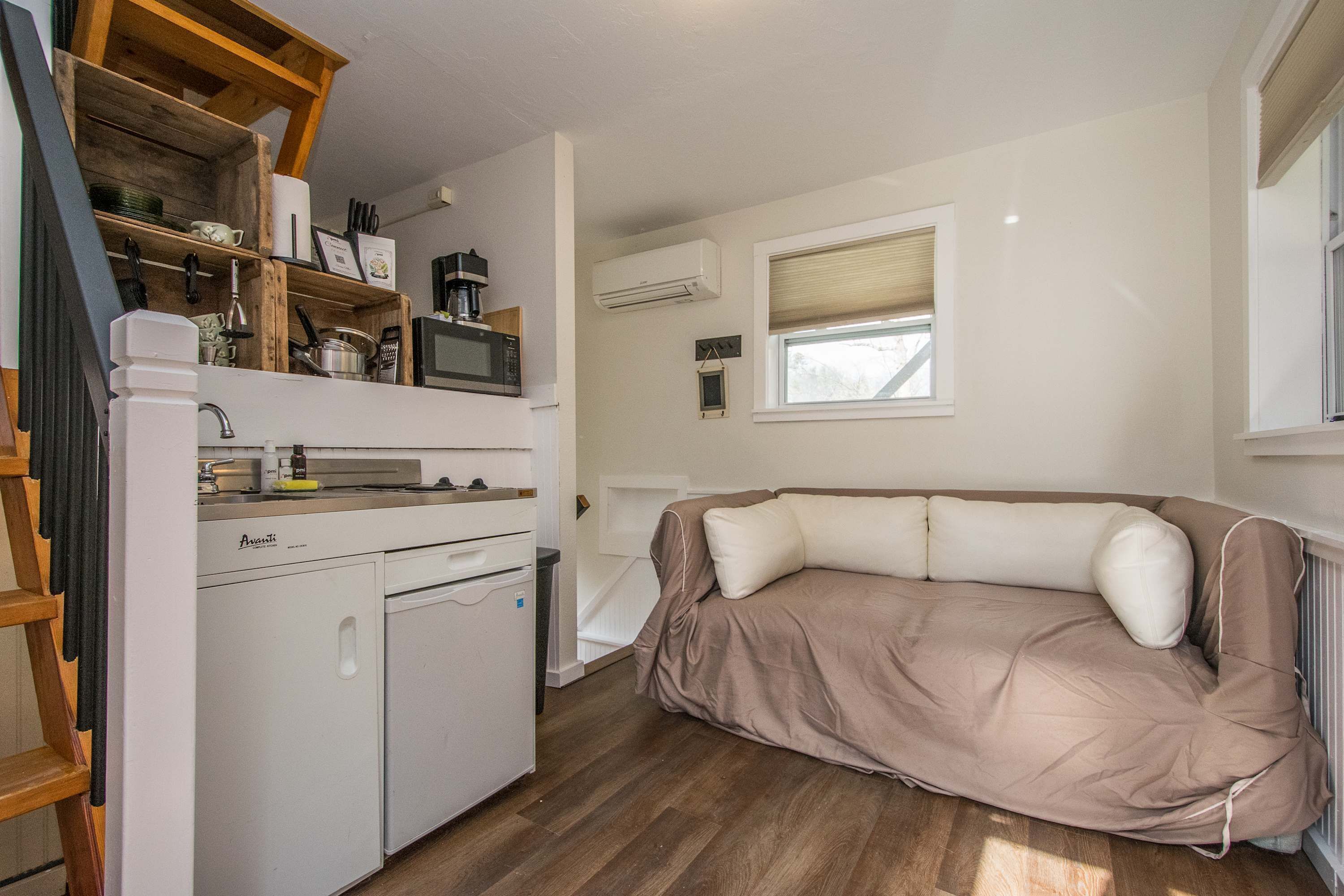 Une cuisine compacte équipée d’un réfrigérateur, d’un four à micro-ondes et d’un évier. Un canapé beige est placé près d’une fenêtre, et une échelle en bois est visible sur la gauche.