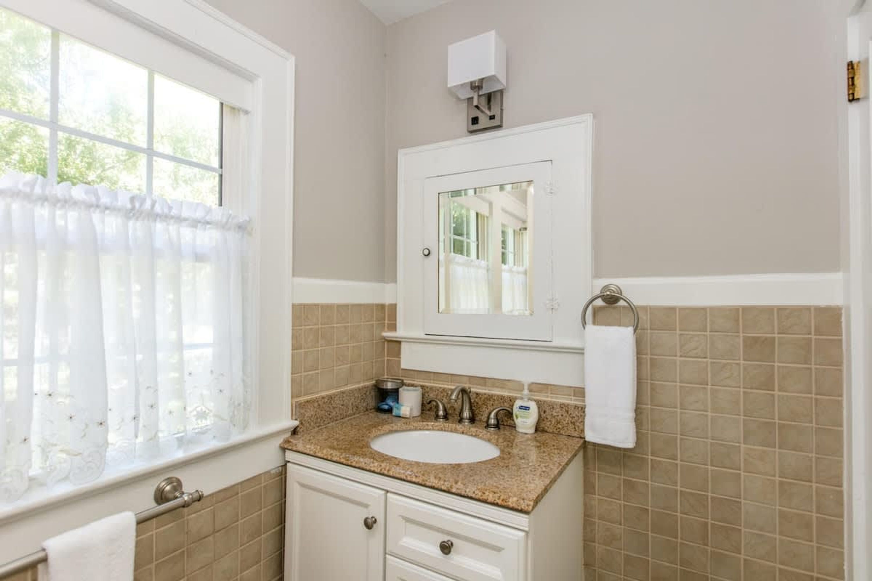 A bathroom with a white sink, beige tiles, a mirror, and a window with sheer curtains.