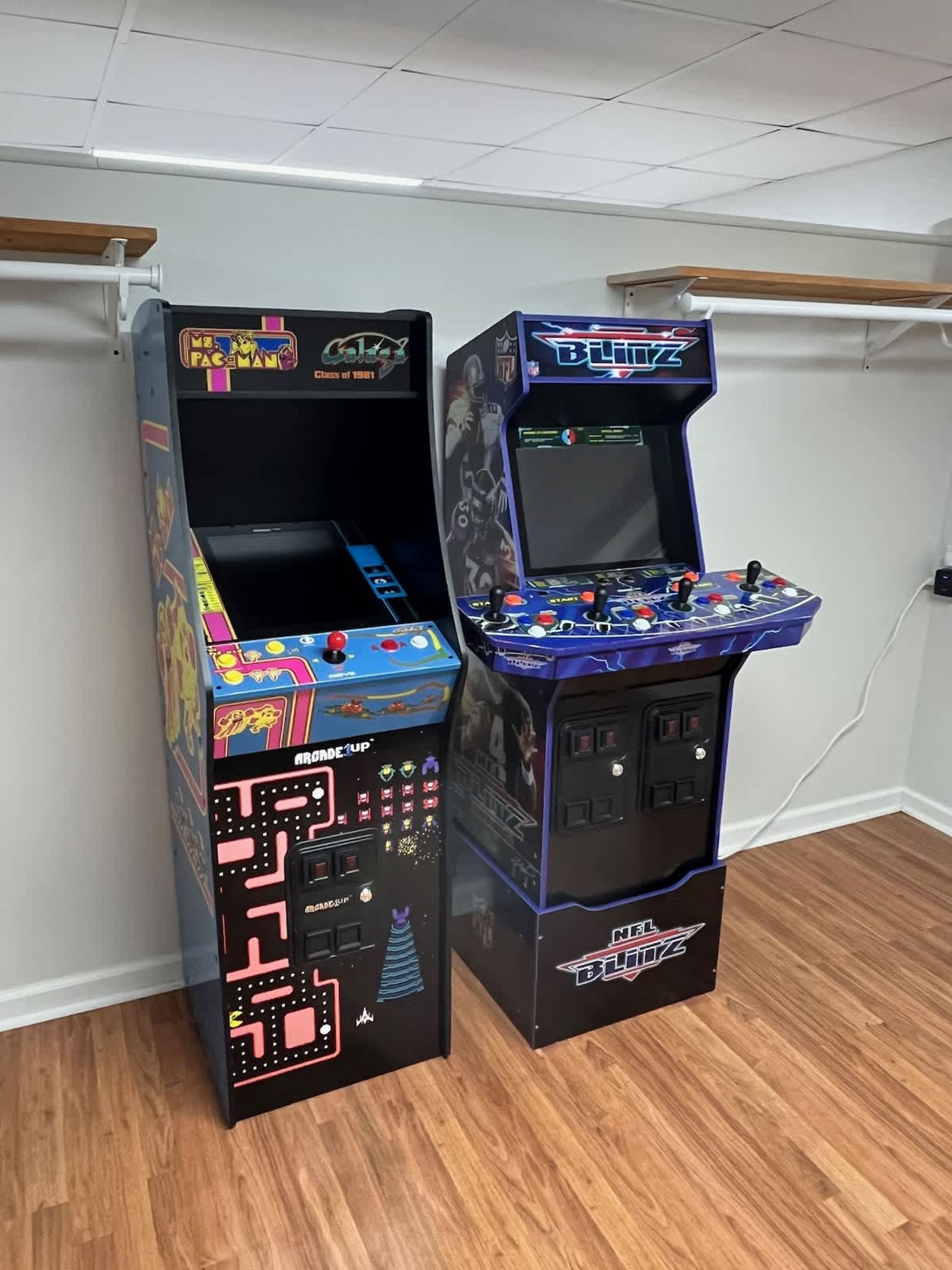 Two arcade machines, one labeled 'Pac-Man' and the other 'NFL Blitz', in a room with wooden flooring and white walls.