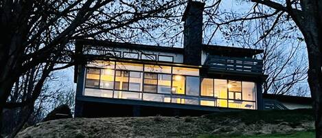 A modern house with a large glass facade, surrounded by trees and a grassy hill.