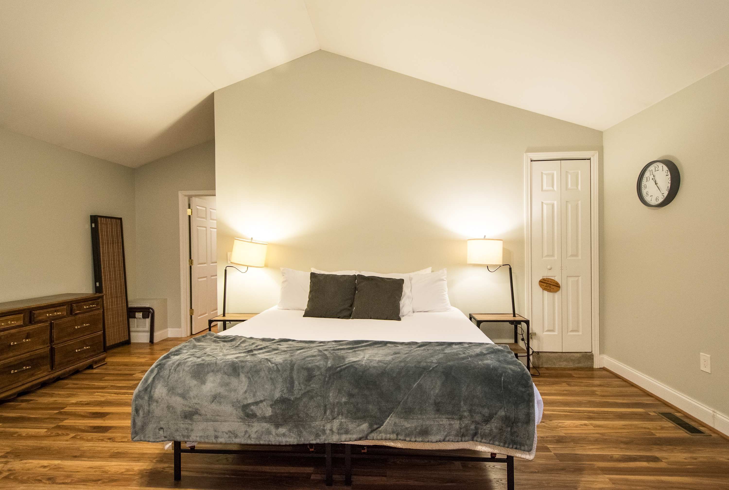 A bedroom with a bed, two bedside lamps, a clock on the wall, and a door.
