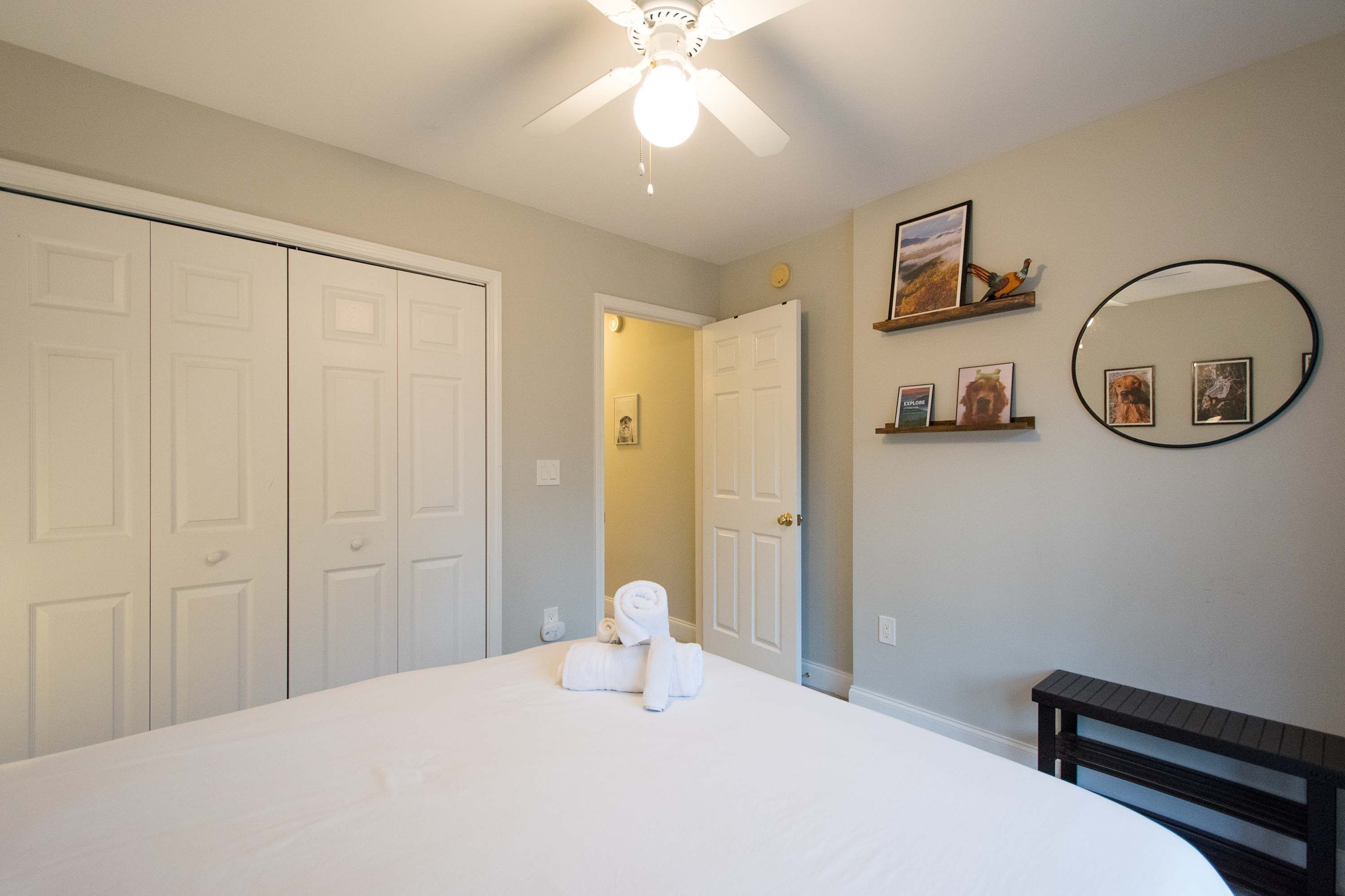 A bedroom with a bed, a ceiling fan, a door, a mirror, and a small shelf with pictures.
