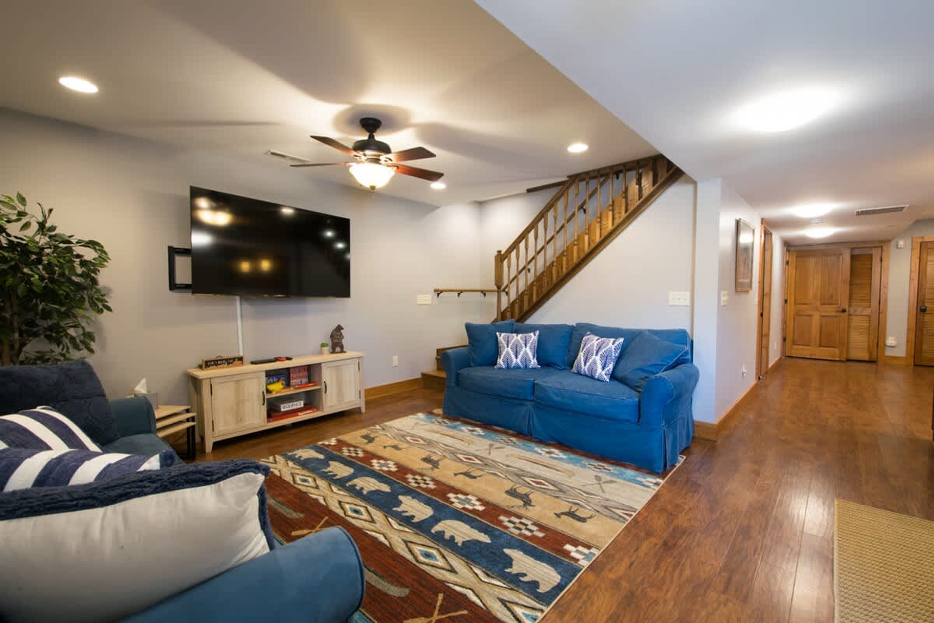 A living room with a blue sofa, a patterned rug, a ceiling fan, and a TV mounted on the wall.