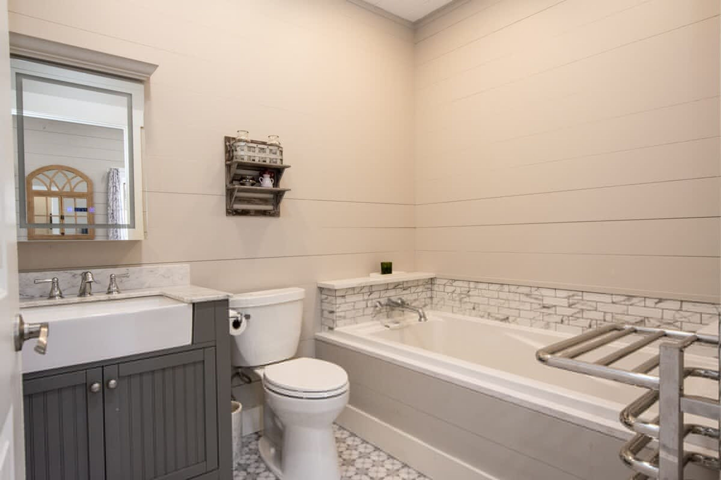 Un baño con lavamanos, inodoro y bañera blancos, con un espejo sobre el lavamanos y un estante con artículos de aseo encima de la bañera.