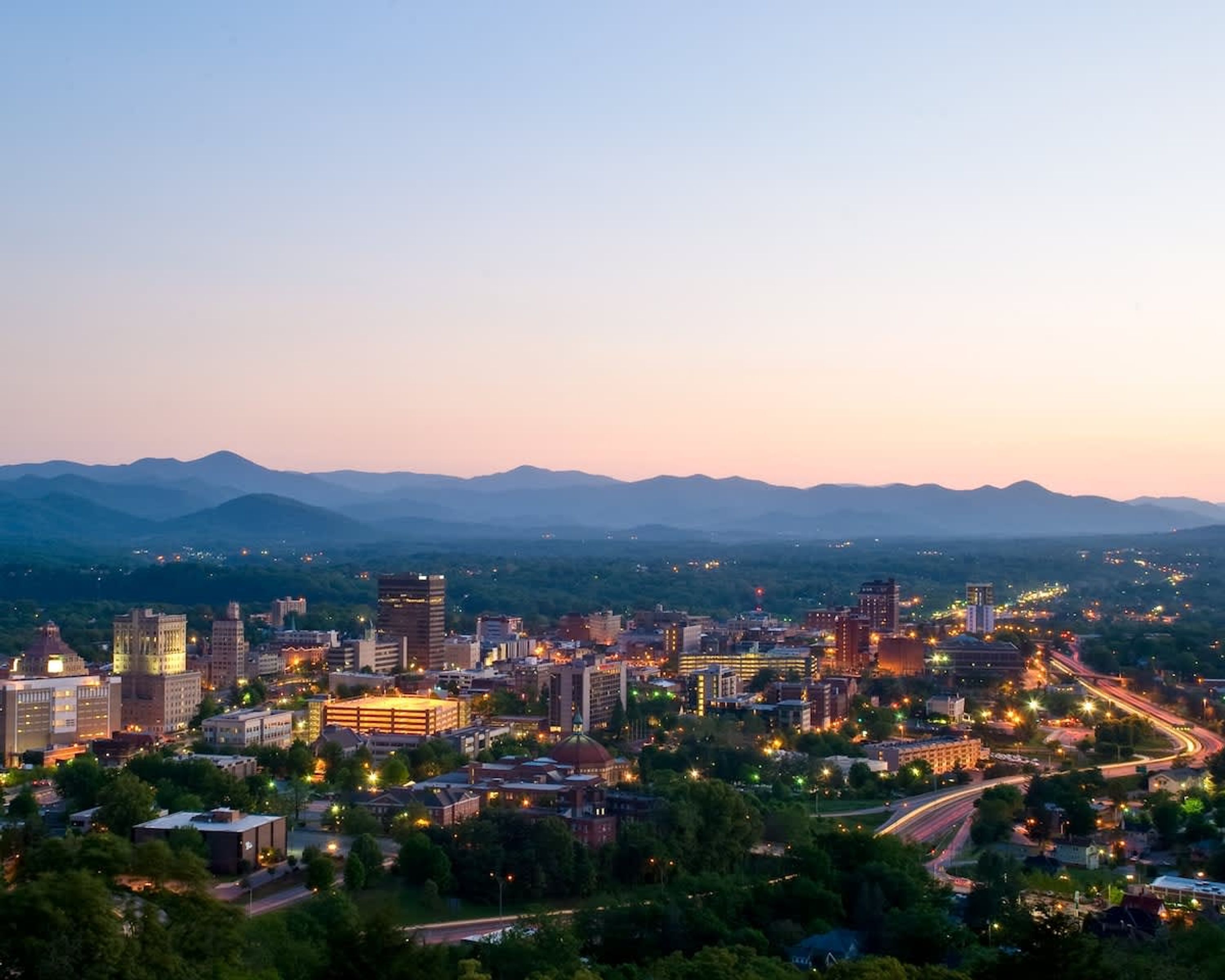 A cityscape at dusk with a mix of modern and historic buildings, a prominent dome structure, and a highway curving through the city.