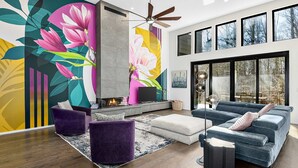 A modern living room with a fireplace, large windows, and a vibrant floral mural. - Luxe 5BR w Home Theater Pool Table Firepit (Alexander)