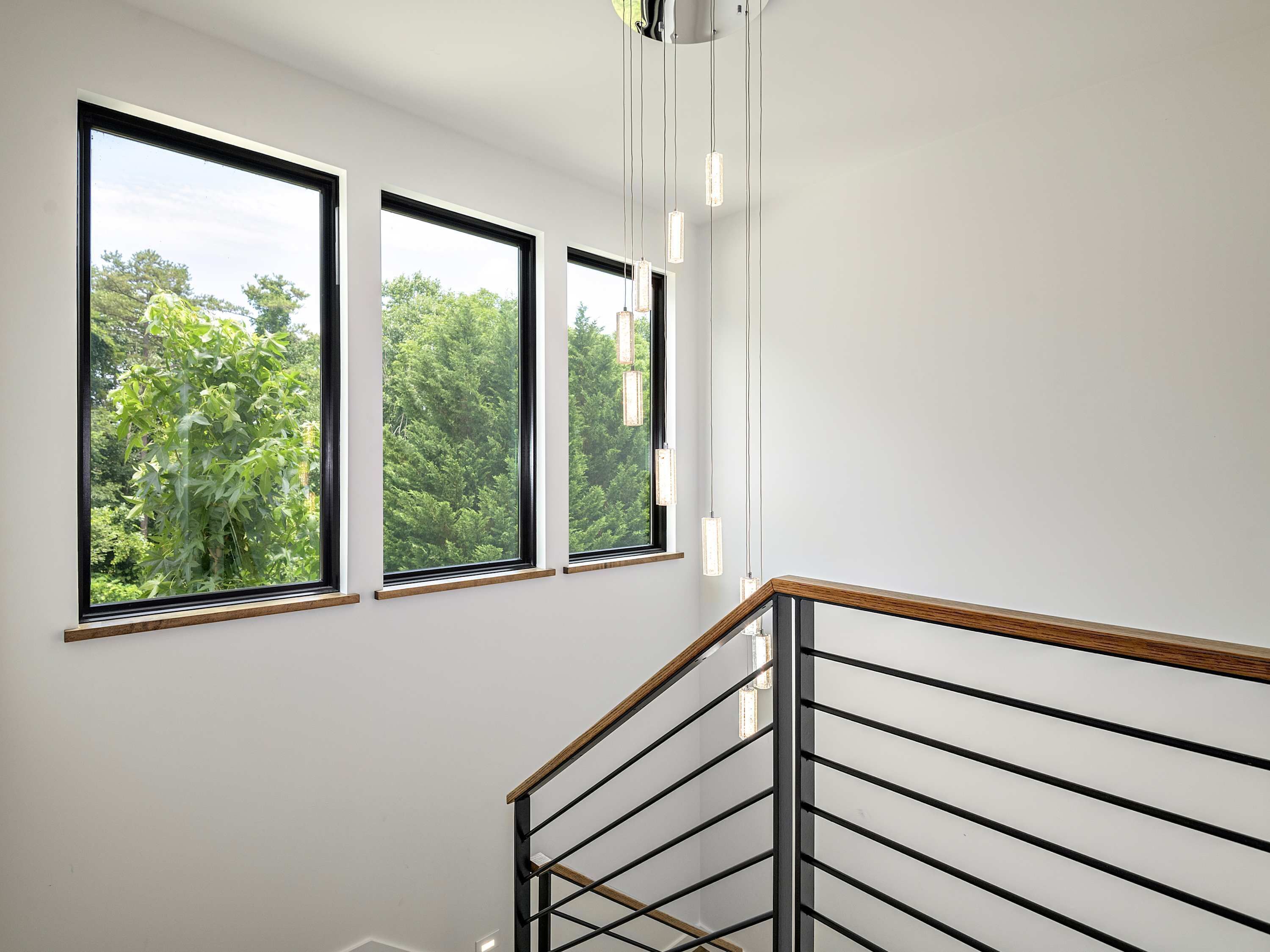 Una escalera con barandilla de madera y balaustradas de metal, que conduce a una plataforma con tres ventanas amplias con vistas a un bosque verde y tupido.