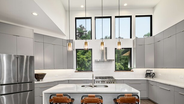 A modern kitchen with grey cabinetry, a stainless steel refrigerator, a central island with stools, and large windows offering a view of greenery. - Luxe 5BR w Home Theater Pool Table Firepit (Alexander)