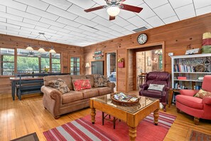 A cozy living room with a wooden ceiling, a ceiling fan, a wooden coffee table, a brown sofa, a red armchair, and a bookshelf. - Lakeside 4BR Mountain Views Firepit Game Room (Lake Lure)