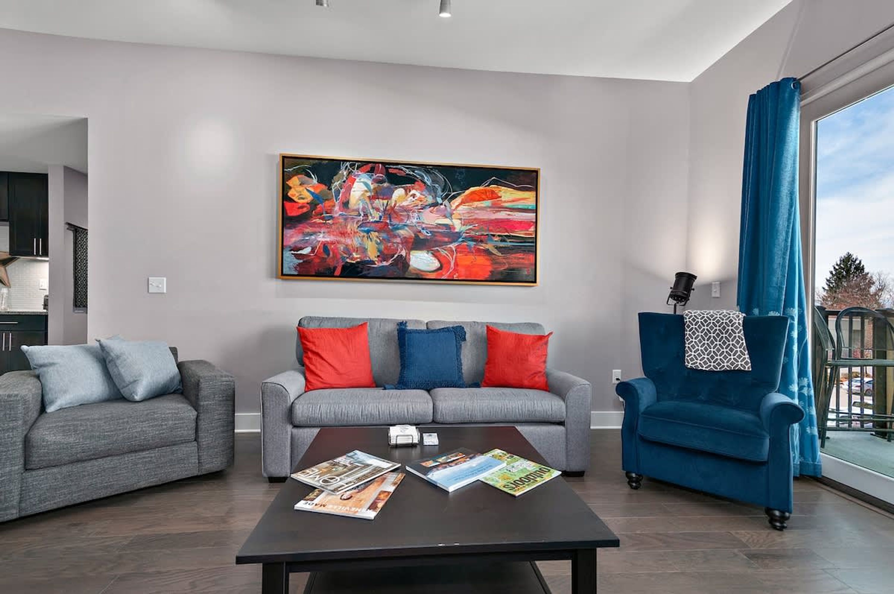 A modern living room with grey sofas, a blue armchair, and a large abstract painting on the wall.