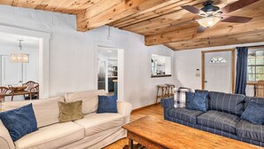A spacious living room with a high wooden ceiling, a ceiling fan, and a large window. There are two sofas, a coffee table, and a dining area with chairs. - Lakefront 4BR w Screened Porch (Lake Lure)
