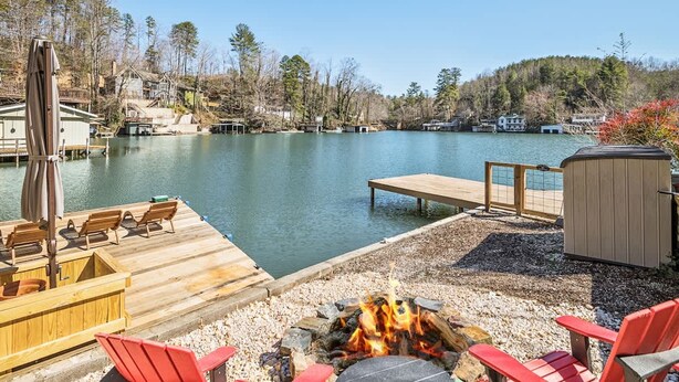 A lakeside deck with wooden chairs, a fire pit, and a wooden dock extending into the water. - Lakefront 4BR w Screened Porch (Lake Lure)