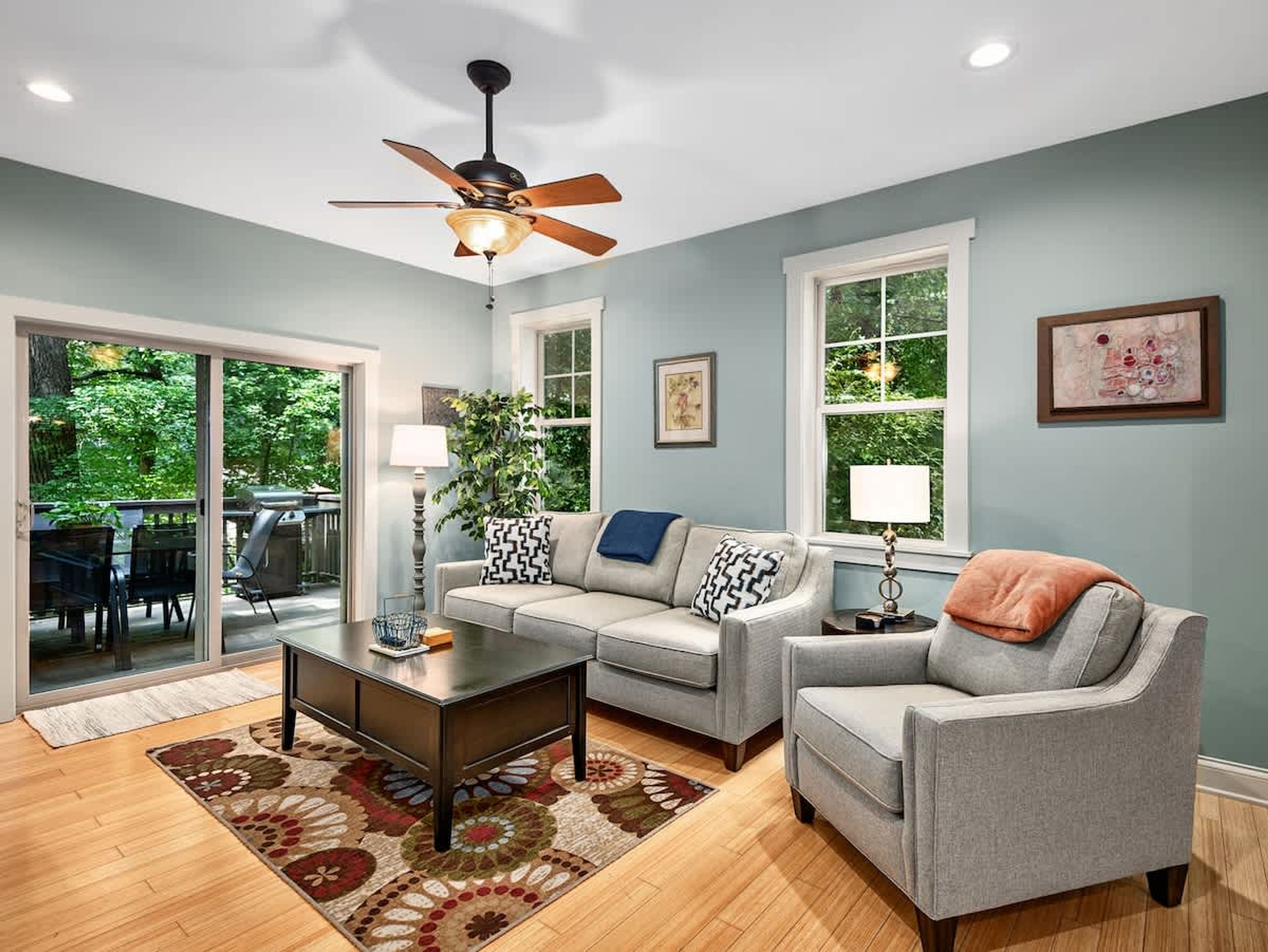 A living room with a sofa, armchair, coffee table, and a ceiling fan. There is a large window with a view of greenery outside.