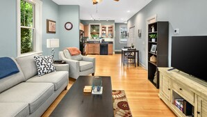 A modern living room with a sofa, coffee table, and TV stand. There is a kitchen in the background with wooden cabinets and a dining area. - Cozy 2BR Townhouse w Deck Walk to W Asheville (Asheville)